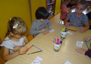 Troje dzieci siedzi przy stoliku i układa z kredek kompozycje według wzorów na karteczkach.
